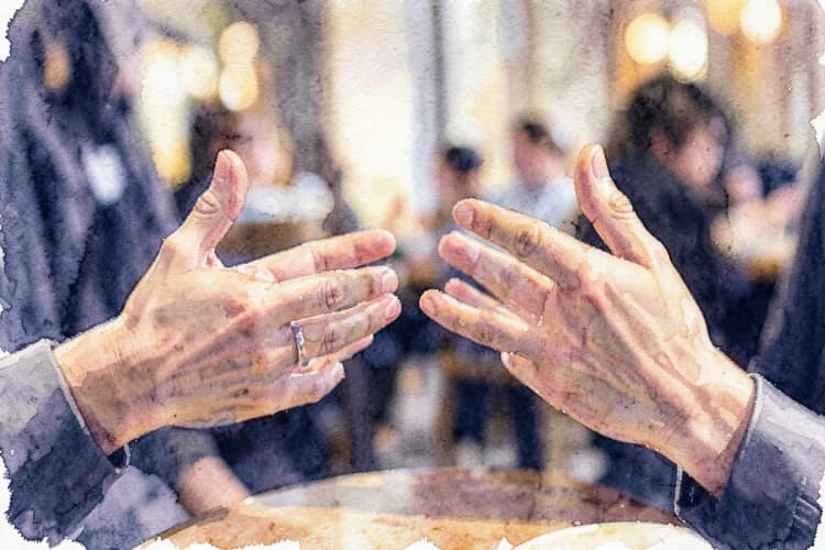 Um close-up de duas mãos gesticulando em uma conversa animada, com um fundo desfocado de um café.